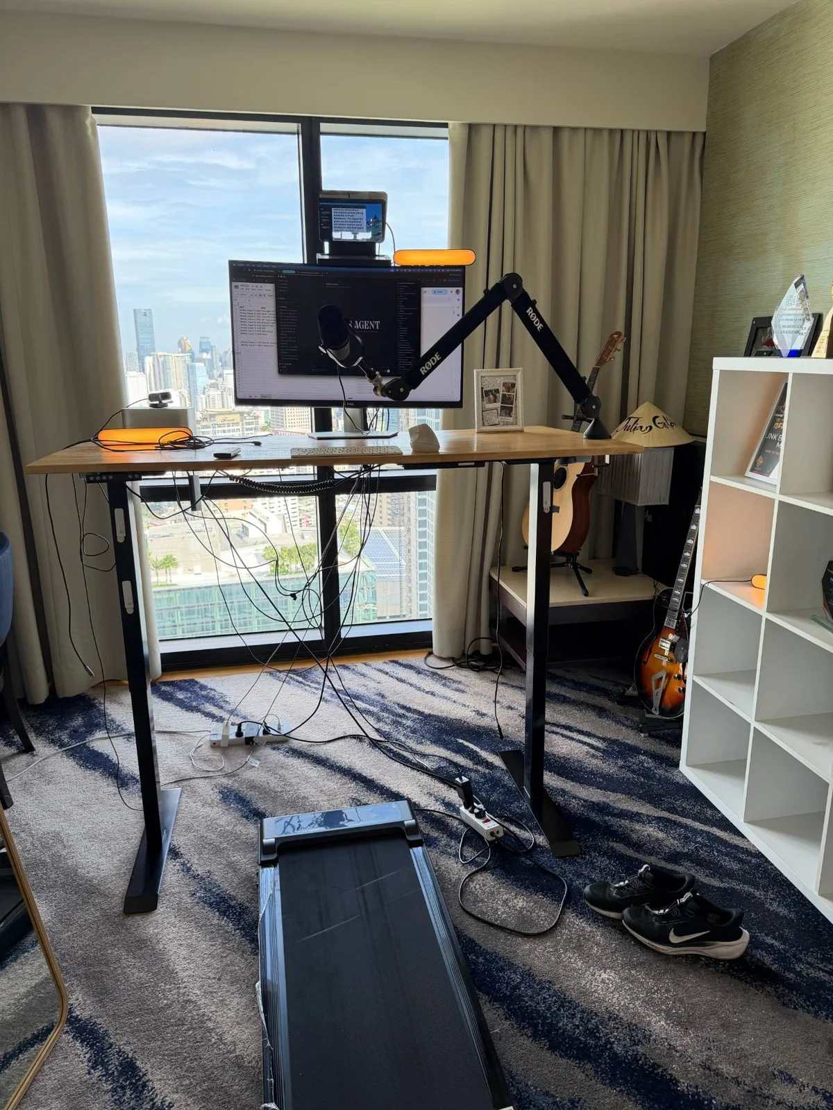 Julian's real desk: a standing desk over a walking treadmill, Rode microphone on a boom arm, monitor with a teleprompter rig on top, and orange key lights